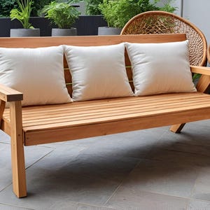 Puede incluir: Un banco de madera con tres cojines blancos. El banco tiene un diseño de listones y está hecho de madera natural. El banco está sentado en un patio.