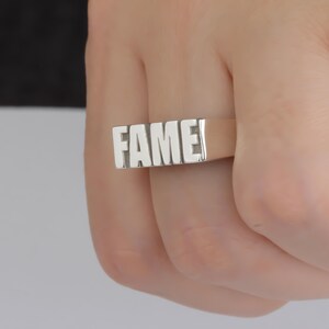 May include: A silver ring with the word "FAME" in white lettering.