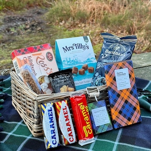 May include: A wicker basket filled with Scottish treats, including chocolate bars, fudge, biscuits, and a box of shortbread fingers. The basket sits on a plaid blanket, with a blurred outdoor background.