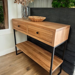 Könnte beinhalten: Ein Konsolentisch aus Holz mit zwei Schubladen und einem unteren Regal. Der Tisch hat eine natürliche Holzoptik und schwarze Metallbeine.