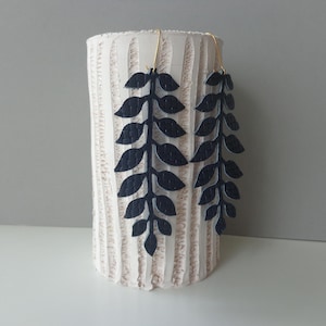 Puede incluir: Un par de pendientes en forma de hojas azul oscuro colgando frente a un jarrón cilíndrico texturizado de color blanquecino. Los pendientes están suspendidos por ganchos dorados. El fondo es gris claro.