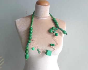 May include: A distinctive green necklace with wooden beads and copper wire. The necklace features a mix of round, oblong, and square beads in various shades of green, creating a modern and artistic look. The necklace is displayed on a mannequin.