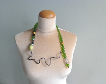 May include: An eye-catching necklace with a black wire base and a mix of green, black, gold, and light blue beads. The necklace is displayed on a mannequin, highlighting its unusual shape and bright colours.