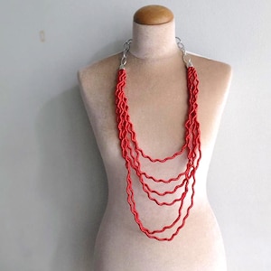 May include: A multi-strand red necklace displayed on a mannequin. The necklace features multiple strands of twisted, rope-like material, connected by silver-colored clasps. The necklace is a bold statement piece, perfect for adding a pop of color.