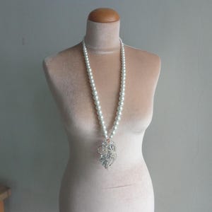 Peut inclure: Un long collier de perles blanches avec un pendentif argenté est présenté sur un mannequin. Le pendentif présente un design détaillé et orné. Les perles sont espacées uniformément.