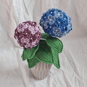 May include: A crocheted hydrangea bouquet in a white pot. The bouquet features two hydrangeas, one purple and one blue, with white beads for accents. The leaves are green.
