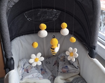 May include: A crocheted baby mobile with a bee, flowers, and yellow and white balls hanging from a black stroller canopy.