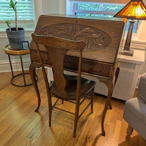 May include: A brown wooden roll-top desk with a carved oval panel on the front and a matching wooden chair with a curved backrest. The desk features a pull-out drawer with metal handles and slender, curved legs, standing on a hardwood floor.