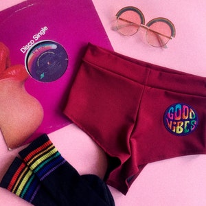 Könnte beinhalten: Burgunderrote, hochgeschnittene Shorts mit einem bunten "Good Vibes"-Patch am rechten Bein. Die Shorts liegen auf einer rosa Oberfläche mit einer Sonnenbrille in Regenbogenfarben, einer schwarz-regenbogenfarbenen Socke und einer Vintage-Schallplatte.