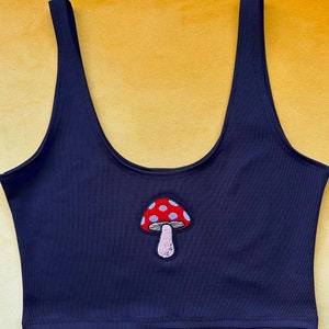 Puede incluir: Un top negro de tirantes con un hongo rojo y blanco bordado en la parte delantera.