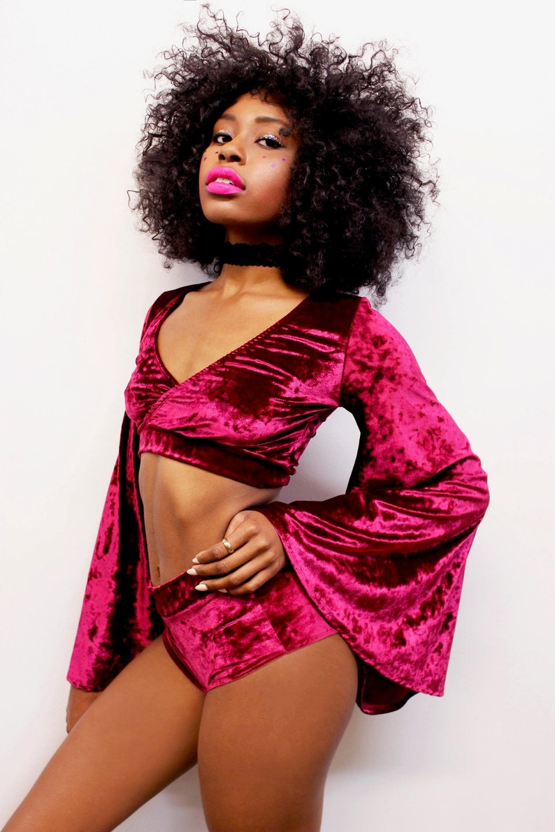 May include: A woman wearing a burgundy velvet crop top and matching shorts. The top has a deep V-neckline and long bell sleeves. The shorts are high-waisted and have a wide leg.