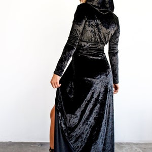 Peut inclure: Une longue robe en velours noir avec une capuche et une fente sur le c&ocirc;t&eacute;. La robe a un corsage ajust&eacute; et une jupe ample. Le mannequin porte la robe avec des bottes &agrave; talons noires.