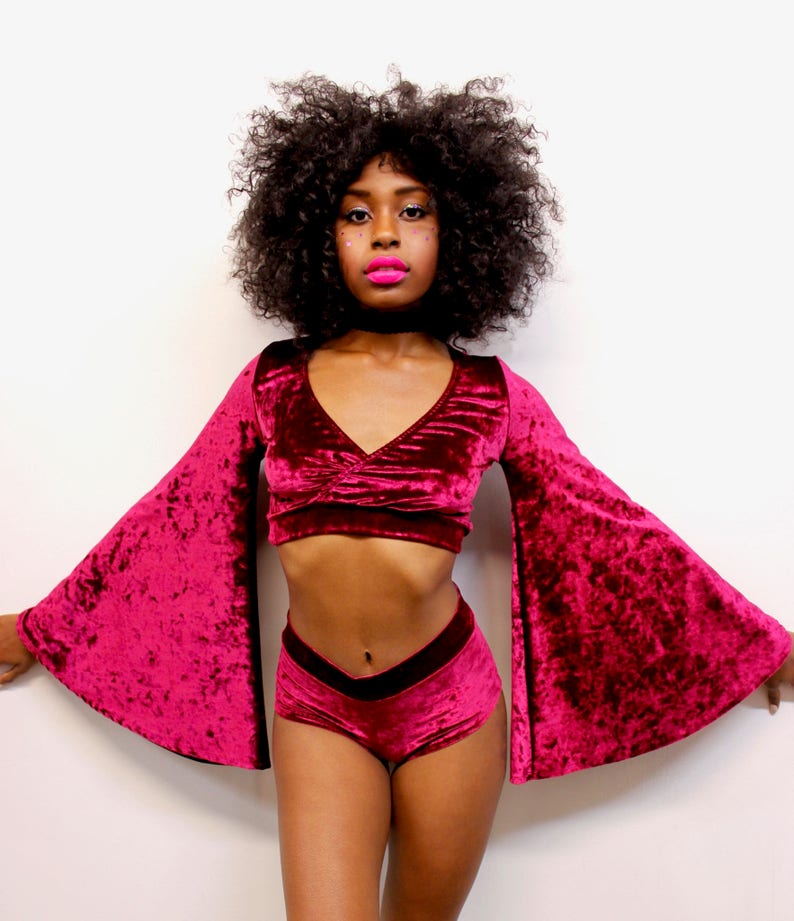 May include: A woman wearing a burgundy velvet crop top with long bell sleeves and matching burgundy velvet shorts. The top has a V-neckline and the shorts have a high waist.