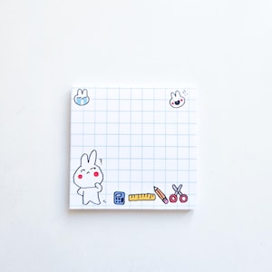Op de afbeelding: Vierkante witte plaknotitie met een blauw rasterpatroon en cartoon konijn illustraties. De notitie bevat een rekenmachine, liniaal, potlood en schaar. Twee konijn illustraties staan in de bovenhoeken.