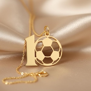 May include: A gold-colored necklace with a soccer ball pendant and the number 1. The soccer ball pendant is a detailed cutout design. The necklace is displayed on a satin surface.
