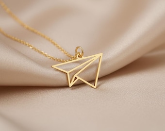 May include: A gold-colored necklace featuring a paper airplane pendant. The pendant is a geometric outline of a paper airplane. The necklace chain and pendant are made of a shiny, reflective metal. The background is a soft, neutral fabric.