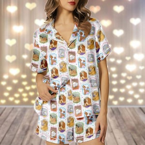 May include: White pajama set with a repeating pattern of framed dog illustrations in various colours. The short-sleeved top and shorts have white piping and a button-down front. The background is blurred with heart-shaped lights.