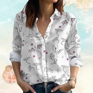 May include: White button-down shirt with a dragon and floral pattern. The long-sleeved shirt features black dragon illustrations with pink accents, moons, stars, and flowers. The shirt is made of a lightweight fabric.