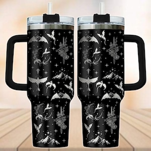 May include: Two black insulated travel mugs with a fantasy design. The design includes silver dragons, swords, moons, and mountains. Each mug has a black handle, a silver rim, and a clear lid with a straw.