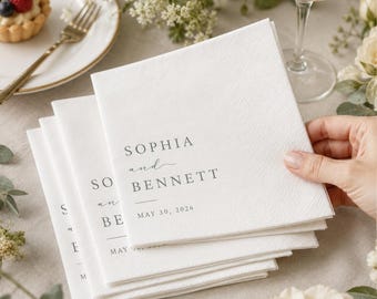 May include: A stack of square white paper napkins featuring black printed text that reads SOPHIA and BENNETT with the date MAY 30, 2026 below. The napkins have a subtle textured finish and are held by a hand, presented in a formal setting.