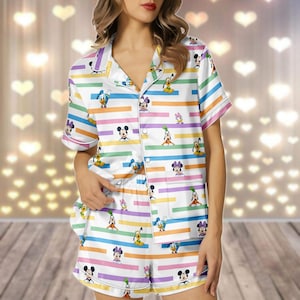May include: White pajama set with a button-down shirt and shorts. The set features a colorful striped pattern with various Disney cartoon characters. The shirt has short sleeves and a collar. The shorts have an elastic waistband.