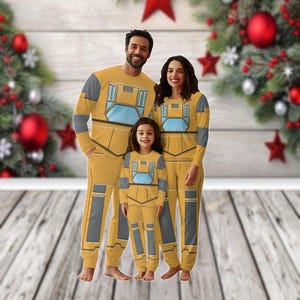 May include: A family of three wearing matching yellow and gray pajamas with a robot design. The pajamas have a long-sleeved shirt and pants. The family is standing in front of a wooden background with a Christmas tree in the background.