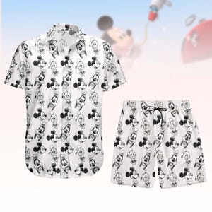 May include: White button-down shirt and matching shorts set. The set features a repeating black and white cartoon character pattern. The shirt has short sleeves and a collar. The shorts have a drawstring waist. The background is a light blue and pink gradient.