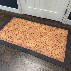 May include: Rectangular doormat with a repeating pattern of floral and diamond shapes in shades of orange, red, and green. The mat has a dark gray border and is placed on a hardwood floor.