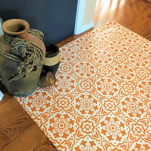 May include: An orange and white patterned rug with a floral design is laid on a hardwood floor. A large, brown ceramic vase with rope detail and a smaller black vase are in the background.