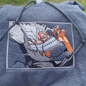 May include: A gray hoodie with a white and orange embroidered image of an anime character with a mask and sharp teeth. The character has white hair and red markings on their face.