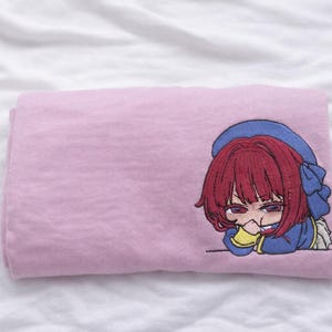 May include: A folded pink textile with an embroidered anime-style character. The character has red hair, a blue bonnet, and a blue outfit. The character is resting their chin on their hands. The textile is on a white, textured surface.