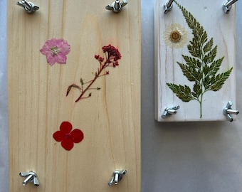 May include: Two rectangular wooden flower presses with metal wing nuts at each corner. The larger press features a light wood grain with a pink flower, a red sprig, and a red four-petaled flower. The smaller press is white with a fern and a white daisy.