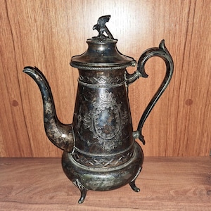 以下が含まれることがあります： 鷲の飾り付きのアンティーク銀メッキコーヒーポット。装飾的な楕円形のカルトゥーシュと湾曲したハンドルを備えた、装飾的なデザインのポットです。注ぎ口があり、3つの小さな足の上に立っています。ヴィンテージアイテムです。