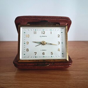 May include: A vintage Europa travel clock in a brown leather case. The clock face is white with gold numerals and hands, and the words "EUROPA" and "2 JEWELS" are printed on the face. The clock is rectangular and has a gold-coloured frame.