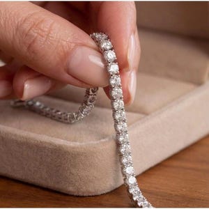 Peut inclure: Un bracelet tennis argenté composé d'une rangée continue de pierres rondes, transparentes et facettées, serties dans une monture métallique. Le bracelet est tenu verticalement, mettant en valeur la taille uniforme et l'éclat de chaque pierre.
