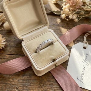Peut inclure: Une alliance éternité argentée ornée d'une rangée de pierres rondes, transparentes et facettées, présentée dans un écrin à bijoux ouvert doublé de velours crème, avec un ruban rose et une étiquette blanche portant l'inscription Forever Yours.