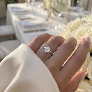 Peut inclure: Une bague argentée ornée d'une pierre ronde transparente montée sur un sertissage à six griffes. L'anneau est lisse et poli, porté au doigt avec une manche en tissu crème visible au premier plan et des roses blanches à proximité.