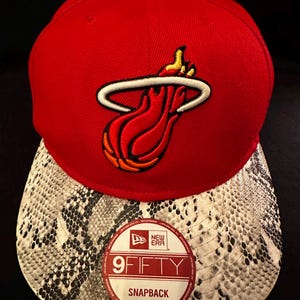 May include: Red and black baseball cap with a Miami Heat logo and a snakeskin patterned brim. The cap has a New Era logo and the text "9FIFTY SNAPBACK".