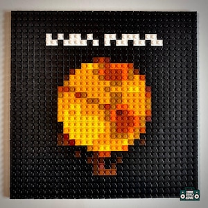 May include: A square black Lego art piece featuring a pixelated image of a yellow and orange object, possibly a mushroom or a flame. The word "M.A.M." is spelled out in white Lego bricks at the top.