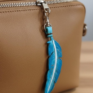 Può includere: Un ciondolo a forma di piuma blu con dettagli bianchi è appeso a un fermaglio argentato attaccato a una borsa marrone. La piuma presenta linee scolpite e piccoli accenti arancioni.