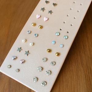 May include: A pink earring display board with various stud earrings. The earrings include small silver stars, hearts, and round studs, along with pearl and rhinestone studs. The board is on a wooden surface.