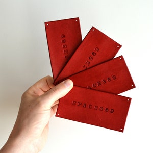 May include: A hand holding a fanned array of four rectangular, red leather tags. Each tag has the embossed text "SPACE369" or "AGE369" or "69SES" or "7369". Each tag has a small hole in the top right corner.