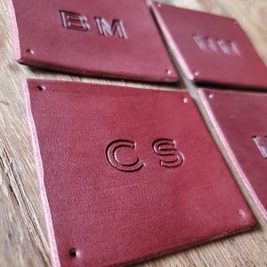 Peut inclure: Quatre dessous de verre carrés en cuir bordeaux avec des initiales en relief. Chaque dessous de verre porte des initiales différentes : BM, MM, CS et HW. Les dessous de verre sont fixés par de petits rivets à chaque coin. Les dessous de verre sont sur une surface en bois.