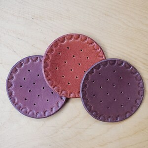 May include: Three round leather coasters in shades of purple and orange. Each coaster has a scalloped edge and small, evenly spaced holes. The coasters are arranged on a light wood surface.