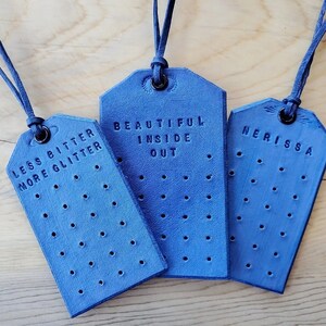 Puede incluir: Tres etiquetas de cuero azul con cordones azul oscuro. Las etiquetas tienen una serie de pequeños agujeros y texto en relieve. Una etiqueta dice "LESS BITTER MORE GLITTER", otra dice "BEAUTIFUL INSIDE OUT" y la tercera dice "NERISSA".