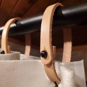 May include: Close-up of a set of natural leather curtain tiebacks. Each tieback is attached to a black metal rod with a black button. The tiebacks are holding up white fabric curtains. The background is a wooden surface.