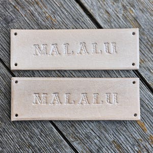 May include: Two rectangular, light tan leather tags, each embossed with the word "MALALU". The tags have rounded corners and two small holes at each end. They are presented on a weathered wooden surface.