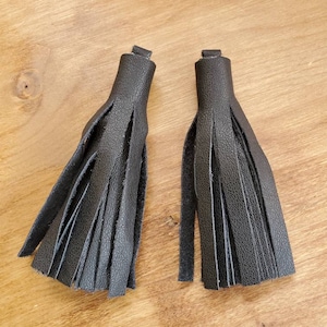 May include: Two black leather tassels, each with a loop at the top and multiple fringed strips. The tassels are displayed on a light brown wooden surface.