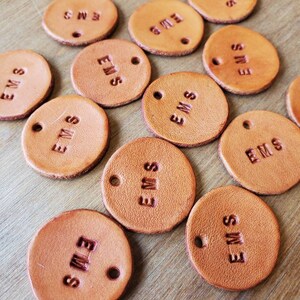 Può includere: Una collezione di etichette rotonde in pelle marrone chiaro con le lettere "EMS" in rilievo. Ogni etichetta ha due piccoli fori e un testo in rilievo leggermente più scuro. Le etichette sono disposte su una superficie di legno marrone chiaro.