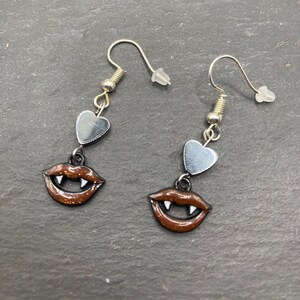 May include: A pair of silver dangle earrings with a brown glitter vampire mouth charm and a silver heart charm.